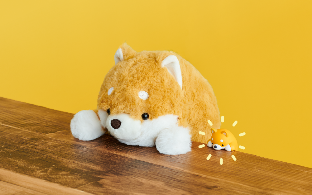 Image is set on a bright yellow background with a wooden table in the foreground. Sitting at the table is the Shiba Inu Posture Pal plush and to the right of it, the Shiba Inu Posture Pal Hipper product with radiating yellow lines around it. 