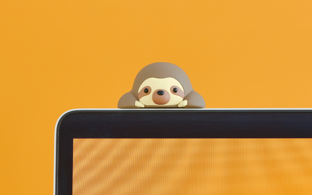 Image, set on a deep yellow background, shows the top edge of a laptop screen with the same color background. Atop the laptop sits the Sloth Posture Pal Hipper. 